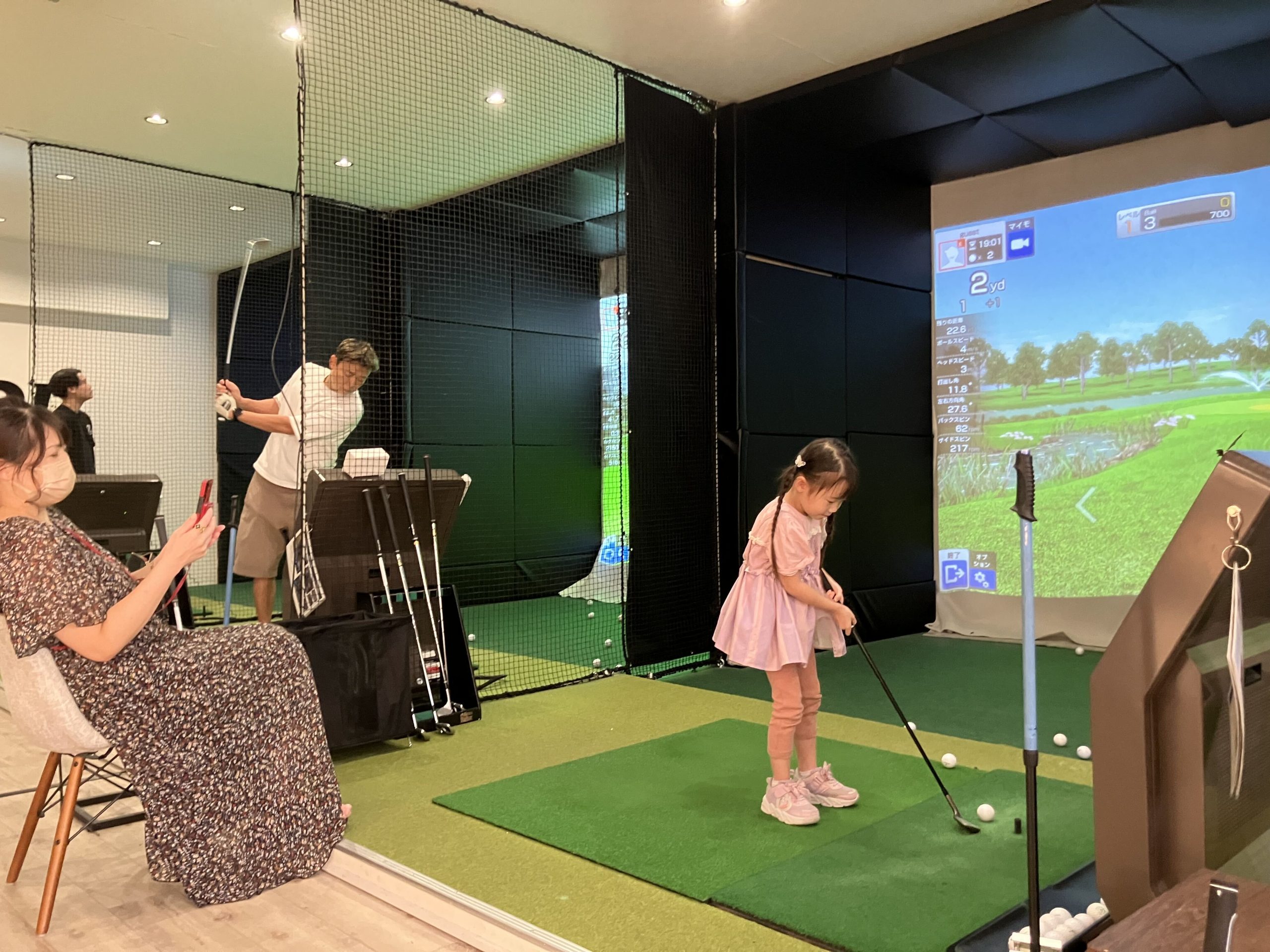 大人がそばで見守る中、シミュレーターの打席でクラブを構えるお子さま